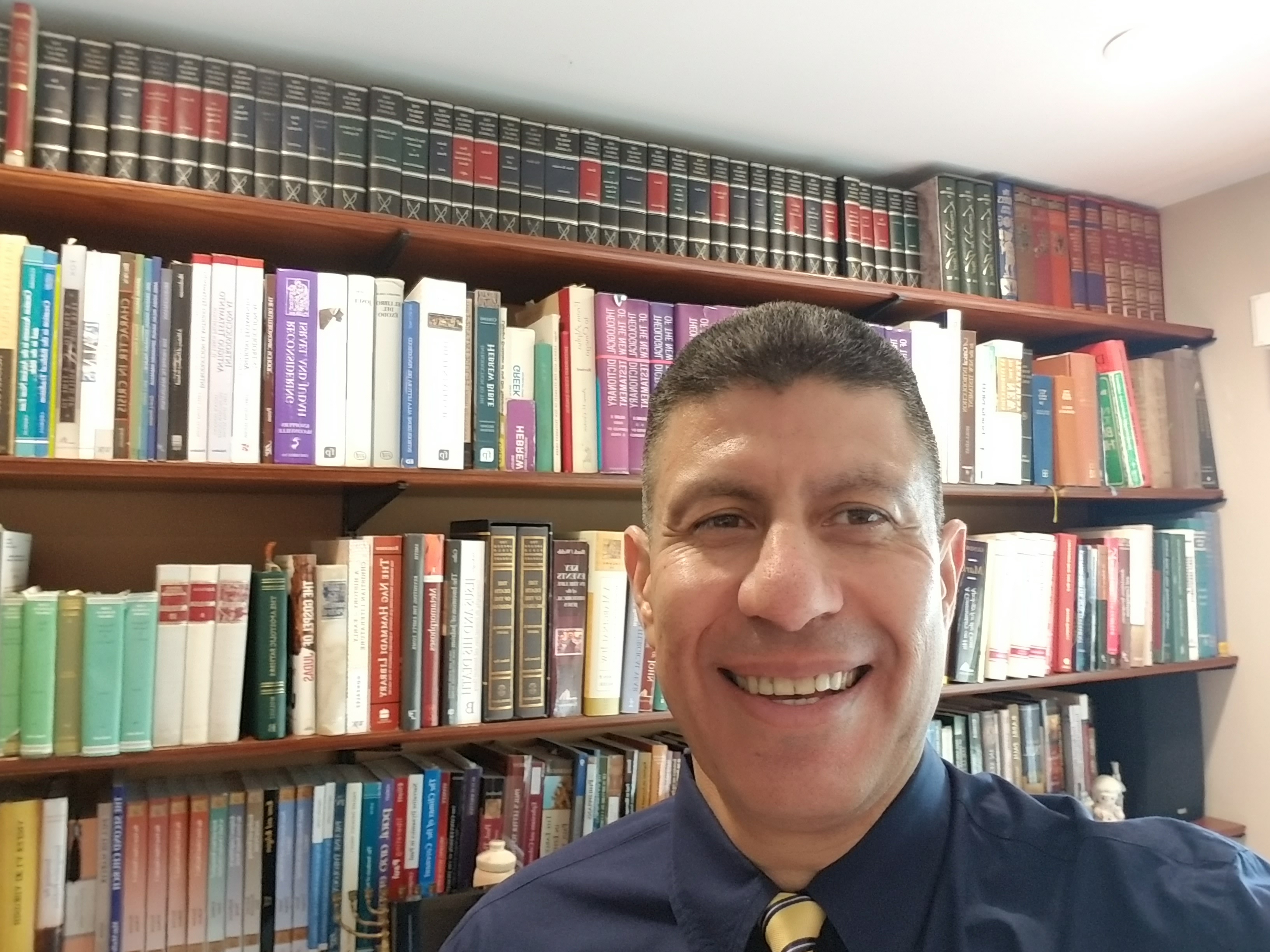 Man smiling in an office, wearing a dress shirt and tie, with shelves of books behind him